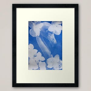 Op de afbeelding: Ingelijste kunstdruk met een blauw-witte flamingo-illustratie. De flamingo staat tegen een blauwe achtergrond, omringd door witte bloemvormen. Het kunstwerk wordt gepresenteerd in een zwart frame met een witte passe-partout.