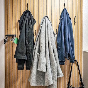 May include: Three coats hanging on hooks on a wood panel wall. The coats are black, grey, and blue. A black leather purse hangs on a hook to the right of the coats.