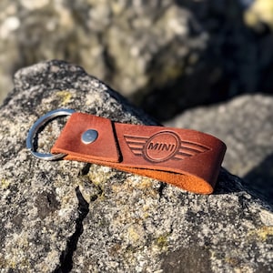 Peut inclure: Porte-clés en cuir marron avec un anneau de clé argenté et un rivet argenté. Le porte-clés est embossé du logo MINI.