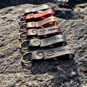 Puede incluir: Varios llaveros de cuero en diferentes colores, incluyendo gris, rojo, marrón y negro. Cada llavero muestra el logotipo y texto de Mazda, con un anillo metálico para llaves. Los llaveros están colocados sobre una superficie rocosa texturizada.