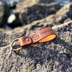 Könnte beinhalten: Ein brauner Leder-Schlüsselanhänger mit dem Mazda-Logo und einem Messingring. Das Logo ist in das Leder geprägt. Das Leder hat eine warme braune Farbe. Der Schlüsselanhänger ist für Schlüssel gedacht.