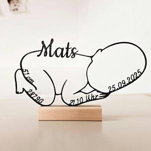 Könnte beinhalten: Eine schwarze glitzernde Silhouette eines Neugeborenen in Fötusstellung auf einem rechteckigen Holzsockel. Das Design zeigt den Namen Mats, die Größe 51 cm, das Gewicht 2879g, die Uhrzeit 21.10 Uhr und das Datum 25.09.2025 in Schreibschrift.