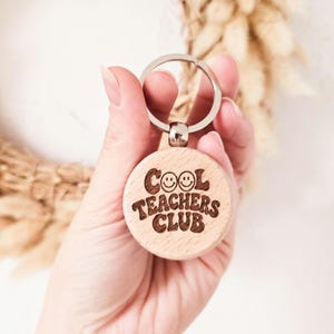 Könnte beinhalten: Ein runder Schlüsselanhänger aus hellem Holz mit einem silbernen Metallring. Die Holzoberfläche ist mit dunkelbraunem Text graviert, der COOL TEACHERS CLUB lautet, wobei zwei kleine Smileys in das Wort COOL integriert sind.