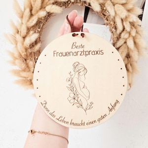 Könnte beinhalten: Ein rundes Schild aus hellem Holz mit einer lasergravierten Illustration einer schwangeren Person und floralen Akzenten. Der Text lautet "Beste Frauenarztpraxis" und "Denn das Leben braucht einen guten Anfang." Es hat ein braunes Lederband zum Aufhängen.