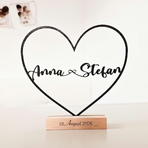 Könnte beinhalten: Schwarze herzförmige Holzdekoration mit den Namen "Anna & Stefan" in Schreibschrift. Auf dem Holzsockel ist das Datum "01. August 2026" eingraviert. Ein romantisches Andenken.