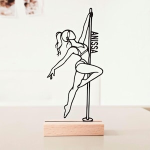 Könnte beinhalten: Eine schwarze Drahtskulptur einer Person, die eine Pole-Dance-Figur ausführt, wobei der Name ANISSA in die vertikale Stange integriert ist. Die Skulptur ist auf einem rechteckigen, hellen Holzsockel mit einem horizontalen Schlitz montiert.