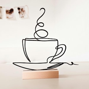 Könnte beinhalten: Eine schwarze Drahtskulptur einer Kaffeetasse mit aufsteigendem Dampf, auf einer Untertasse und einem kleinen Holzsockel. Das minimalistische Design zeigt eine durchgehende Linie, die Tasse, Henkel und Dampf bildet.
