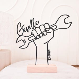 Könnte beinhalten: Schwarzes Metallkunstwerk, das eine Hand zeigt, die einen Schraubenschlüssel hält, montiert auf einem hellen Holzsockel. Der Name "Geselle" ist in Kursivschrift über dem Schraubenschlüssel geschrieben, mit "2024" auf dem Schraubenschlüssel. Der Name "Maurice" steht vertikal auf dem Sockel.