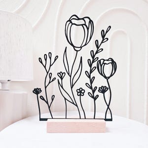 Könnte beinhalten: Schwarze Metall-Blumenskulptur mit stilisierten Blumen und Blättern, montiert auf einem hellrosa Holzsockel. Das Design umfasst Tulpen und andere florale Elemente, ideal für die Inneneinrichtung.