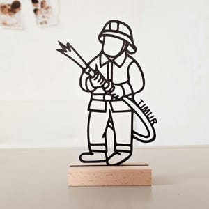 Könnte beinhalten: Eine schwarze Scherenschnitt-Figur eines Feuerwehrmanns mit Helm und Uniform, der einen Schlauch hält, auf dem der Name TIMUR steht. Die Figur ist auf einem rechteckigen, hellen Holzsockel montiert und steht aufrecht als Dekoration.