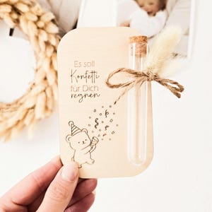 Könnte beinhalten: Eine rechteckige Holzkarte mit abgerundeten Ecken, bedruckt mit dem Text Es soll Konfetti für Dich regnen und einer Strichzeichnung eines Bären mit Partyhut. Ein klares Glas-Reagenzglas mit Korken ist mit einer Juteschnur und getrocknetem Gras befestigt.