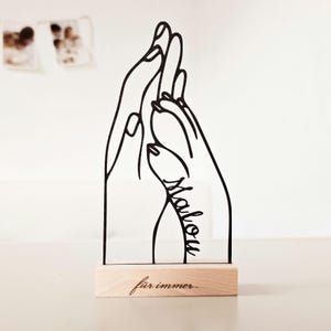Könnte beinhalten: Schwarze Metallskulptur, die zwei Hände zeigt, die sich berühren, auf einem hellen Holzsockel. Der Name "Malou" ist in Schreibschrift auf einer Hand geschrieben, und "für immer" ist auf dem Sockel eingraviert.