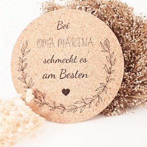 Könnte beinhalten: Ein runder Korkuntersetzer mit dem Text "Bei Oma Marina schmeckt es am Besten" in schwarzer Schrift, umrahmt von einem Blattdesign. Ein kleines Herz befindet sich unter dem Text. Der Untersetzer ist von Trockenblumen umgeben.