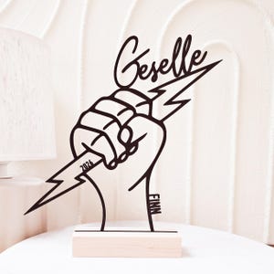 Könnte beinhalten: Schwarze Metallskulptur mit einer geballten Faust, die einen Blitz hält. Der Name "Geselle" steht in Schreibschrift über der Faust. Das Jahr "2026" steht auf dem Blitz, und der Name "FINN" steht auf dem Sockel.