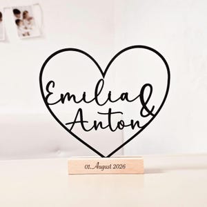 Könnte beinhalten: Ein schwarzer herzförmiger Ausschnitt mit den Namen Emilia & Anton in Schreibschrift, montiert auf einem rechteckigen hellen Holzsockel mit dem eingravierten Datum 01. August 2026, das mittig auf der Vorderseite des Holzblocks steht.