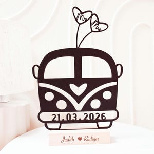 Könnte beinhalten: Ein schwarzer Hochzeitstortenaufsatz aus Holz mit einem stilisierten Bus und zwei herzförmigen Verzierungen. Das Datum "21.03.2026" ist zusammen mit den Namen "Judith" und "Rüdiger" auf einem Holzsockel abgebildet. Der Aufsatz ist für eine Hochzeit bestimmt.