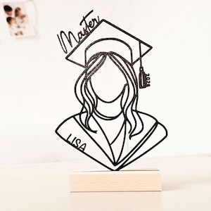 Könnte beinhalten: Ein schwarzer, glitzernder Tortenaufleger in Silhouettenform, der eine graduierte Person mit Doktorhut zeigt, beschriftet mit Master und 2026, sowie dem Namen LISA auf dem Talar, montiert auf einem rechteckigen, hellen Holzsockel mit langem Haar.