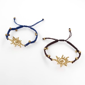 Op de afbeelding: Twee handgemaakte armbanden. Eén is marineblauw met goudkleurige kralen en een goudkleurige zon-bedel. De andere is bruin met goudkleurige kralen en een goudkleurige zon-bedel. Beide armbanden staan op een witte achtergrond.