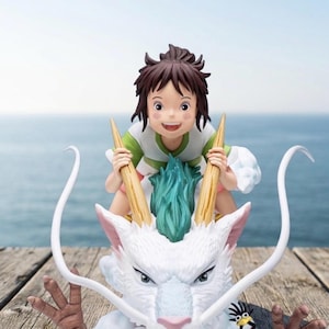 May include: A figurine of a young girl riding a white dragon with a blue and green mane. The girl is wearing a green and white shirt and has brown hair. The dragon has long, curved horns and a stern expression. The background is a blurred view of the ocean.