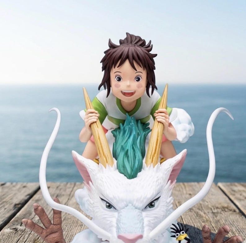 May include: A figurine of a young girl riding a white dragon with a blue and green mane. The girl is wearing a green and white shirt and has brown hair. The dragon has long, curved horns and a stern expression. The background is a blurred view of the ocean.