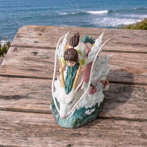 May include: A detailed figurine depicting a scene from a popular animated film. The sculpture features a young girl riding a white dragon through the clouds. The dragon has a long, serpentine body and is surrounded by fluffy white clouds. The figurine is set against a backdrop of the ocean.