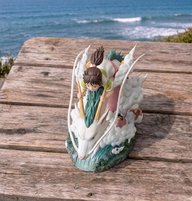 May include: A detailed figurine depicting a scene from a popular animated film. The sculpture features a young girl riding a white dragon through the clouds. The dragon has a long, serpentine body and is surrounded by fluffy white clouds. The figurine is set against a backdrop of the ocean.