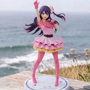 May include: A figurine of a female anime character in a pink dress with a ruffled top and layered skirt. She has purple hair, pink gloves, and pink tights. The figure is holding a microphone and has one arm raised. The base is white with the text "Trio-Try-IT Figure."