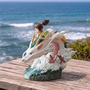 Pode incluir: Estatueta detalhada que retrata uma jovem montada em um dragão branco através das nuvens. O dragão tem uma barriga rosa e é cercado por nuvens brancas fofas. A menina usa uma roupa verde e branca e segura varas de madeira. O fundo mostra o mar.