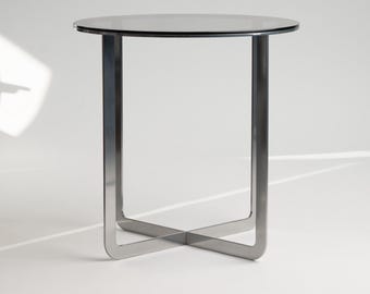 Puede incluir: Una mesa auxiliar moderna con una tapa redonda de cristal oscuro y una base de metal cepillado. La base presenta un diseño cruzado único para mayor estabilidad. La mesa es adecuada para salones o dormitorios. Altura: aprox. 50 cm.