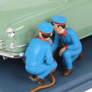 Könnte beinhalten: Eine Figur, die zwei Mechaniker in blauen Overalls und Mützen zeigt, die an einem hellgrünen Oldtimer arbeiten. Die Mechaniker knien und scheinen einen Schlauch zu inspizieren. Die Szene spielt auf einer blauen Oberfläche.