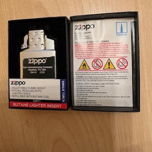 May include: A silver Zippo butane lighter insert in a black box with a white instruction manual. The insert features the Zippo logo and text. The manual includes safety warnings, icons, and the text Zippo Torch. The box label reads Single Torch.