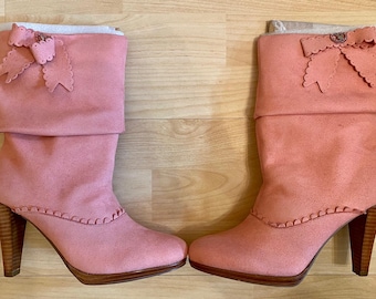 May include: A pair of pink suede boots with a high heel and a decorative bow. The boots have a folded-over cuff and a wooden heel. The heel height is approximately 3 inches.