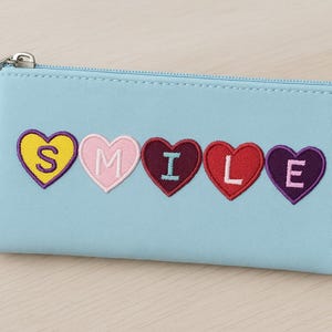 Może przedstawiać: Jasnoniebieska kosmetyczka zapinana na zamek z napisem "SMILE" wyszytym w kolorowych kształtach serc. Serca są żółte, różowe, bordowe, czerwone i fioletowe. Kosmetyczka ma srebrny suwak.