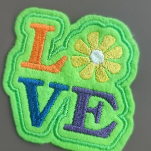 May include: Embroidered patch with the word "LOVE" in orange, blue, and purple, and a yellow flower with a white center. The patch has a green, fuzzy border and is on a gray background.