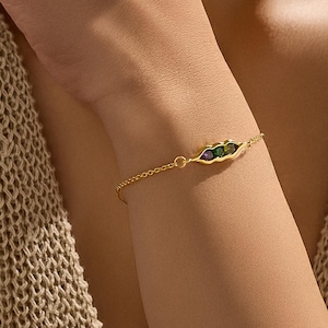 Pode incluir: Uma delicada pulseira dourada com um design de vagem de ervilha com três pedras preciosas coloridas. A pulseira é usada no braço de uma pessoa, mostrando sua corrente fina e as cores vibrantes das pedras. É uma joia.