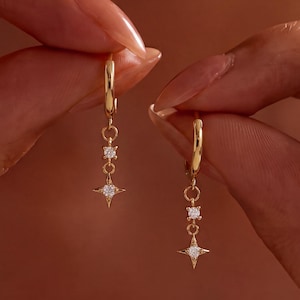 May include: A pair of gold hoop earrings featuring a dangling charm design. Each earring consists of a small polished gold hoop connected to a vertical chain link that holds a single round clear stone and a four-pointed star charm with a centre stone.