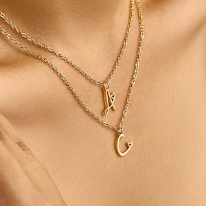 Könnte beinhalten: Goldfarbene, mehrreihige Halskette mit zwei Ketten. Die obere Kette hat einen Anhänger mit dem Buchstaben "A", die untere Kette einen Anhänger mit "G". Die Halskette wird um den Hals getragen.