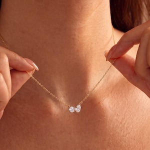 Könnte beinhalten: Eine zarte goldfarbene Kettenhalskette mit einem kleinen Anhänger, der aus zwei klaren, facettierten Steinen in Schmetterlingsform besteht. Die Halskette liegt auf dem Schlüsselbein und zeigt ein Design aus einer dünnen, polierten Metallgliederkette.