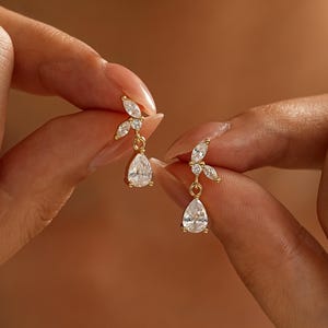 May include: A pair of gold-toned drop earrings featuring a cluster of clear, marquise-cut stones at the top and a single, larger pear-shaped clear stone dangling from the base. The metal setting is polished gold, providing a bright, reflective finish.