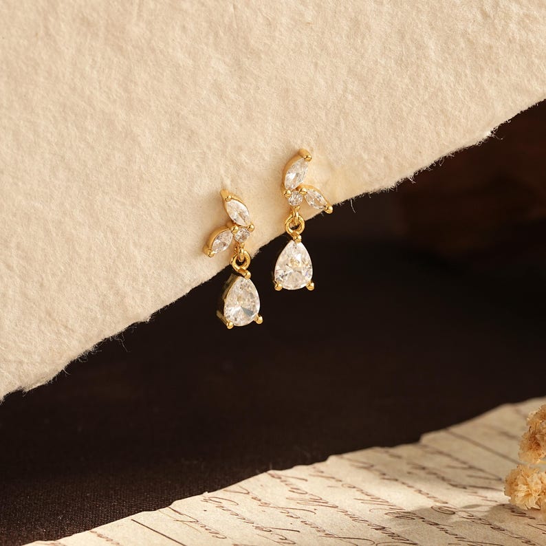 May include: A pair of gold-toned drop earrings featuring a cluster of clear marquise-cut stones at the top and a single clear pear-shaped stone dangling from the base. The earrings are designed with a delicate metal setting and clear crystals.