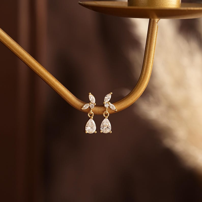 May include: A pair of gold-toned earrings featuring a delicate leaf-like design set with small clear marquise-cut stones, with a single dangling pear-shaped clear stone suspended from the base of each earring, resting on a curved gold metal arm.