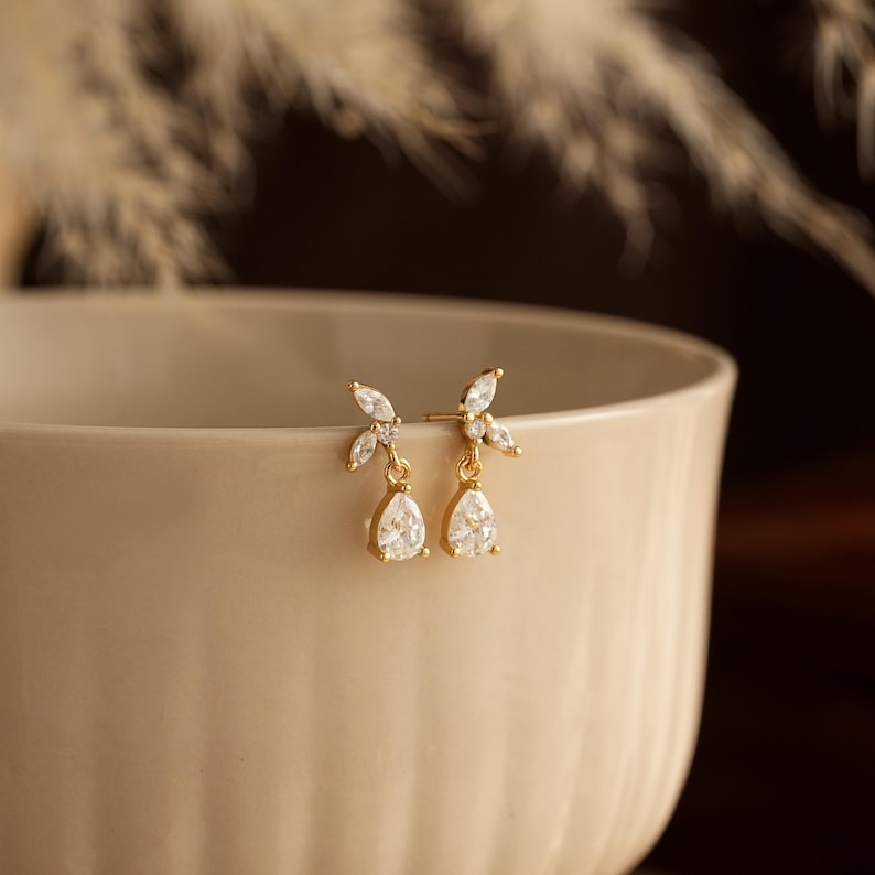 May include: A pair of gold-tone stud earrings featuring a cluster of three marquise-cut clear stones arranged in a leaf-like pattern, with a single pear-shaped clear stone dangling from the base of each earring, set against a light beige bowl.