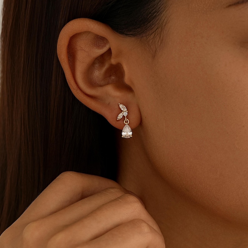 May include: A gold-toned earring featuring two small marquise-cut clear stones arranged in a leaf-like pattern at the top, with a single dangling pear-shaped clear stone suspended below, all set in a delicate metal frame worn on the earlobe.