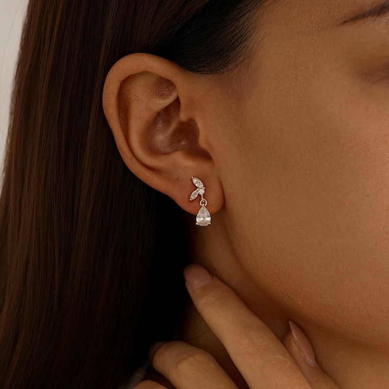 May include: A gold-toned earring featuring two small marquise-cut clear stones arranged in a leaf-like pattern at the top, with a single dangling pear-cut clear stone suspended below. The metal setting is polished and reflective in the light.