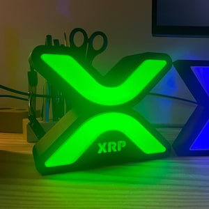 May include: A black and green illuminated sign with the word "XRP" in white letters. The sign is shaped like an X and is lit up with green light.