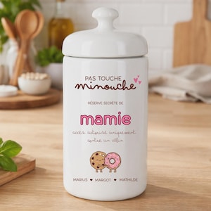 Peut inclure: Un pot en céramique blanc avec un couvercle, avec du texte en français et des illustrations. Le pot indique "PAS TOUCHE minouche" et "RÉSERVE SECRÈTE DE mamie". En dessous, des illustrations de deux beignets et des noms: MARIUS, MARGOT et MATHILDE.
