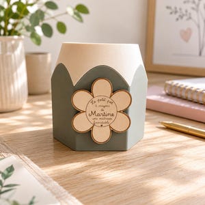 Peut inclure: Un pot à crayons hexagonal avec une base vert sauge et un sommet en bois clair. Une plaque en bois en forme de fleur est fixée sur le devant, portant le texte gravé : Le petit pot à crayons de Martine une maîtresse formidable.