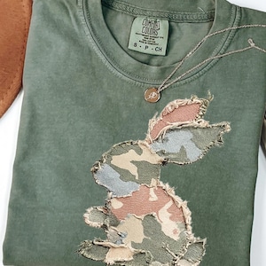 May include: Moss green t-shirt with a camouflage bunny applique. The shirt has a crew neck and short sleeves. A gold-colored necklace with a pendant is draped on the shirt. The tag reads "Comfort Colors".