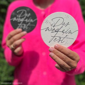 Könnte beinhalten: Zwei runde Untersetzer, einer schwarz und einer weiß, jeweils mit dem handgeschriebenen Satz "Das wird ein Fest." Die Untersetzer werden vor einem leuchtend pinkfarbenen Hintergrund gehalten, mit dem Text in einer fließenden Schrift.