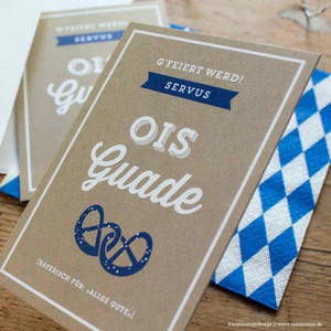 Könnte beinhalten: Zwei braune Karten mit weißen Rändern und Text. Die Karten tragen die Worte "OIS Guade" in großen weißen Buchstaben, mit einem blauen Brezel-Design. Der Text "G'FEIERT WERD! SERVUS" steht oben. Eine blau-weiß karierte Serviette liegt daneben.